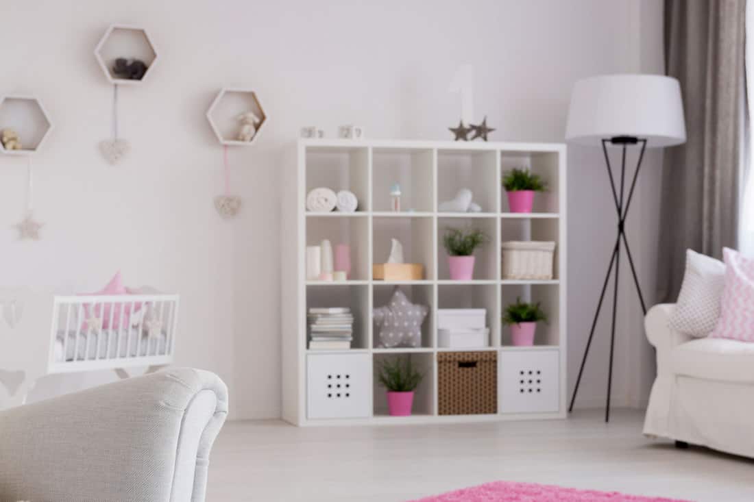 Baby girl room in pastel colors with a teddy bear sittin on an armchair with the comfortable couch, book case, cradle and a pink carpet in the background, How To Organize Clothes Without A Dresser [6 Great Ways]
