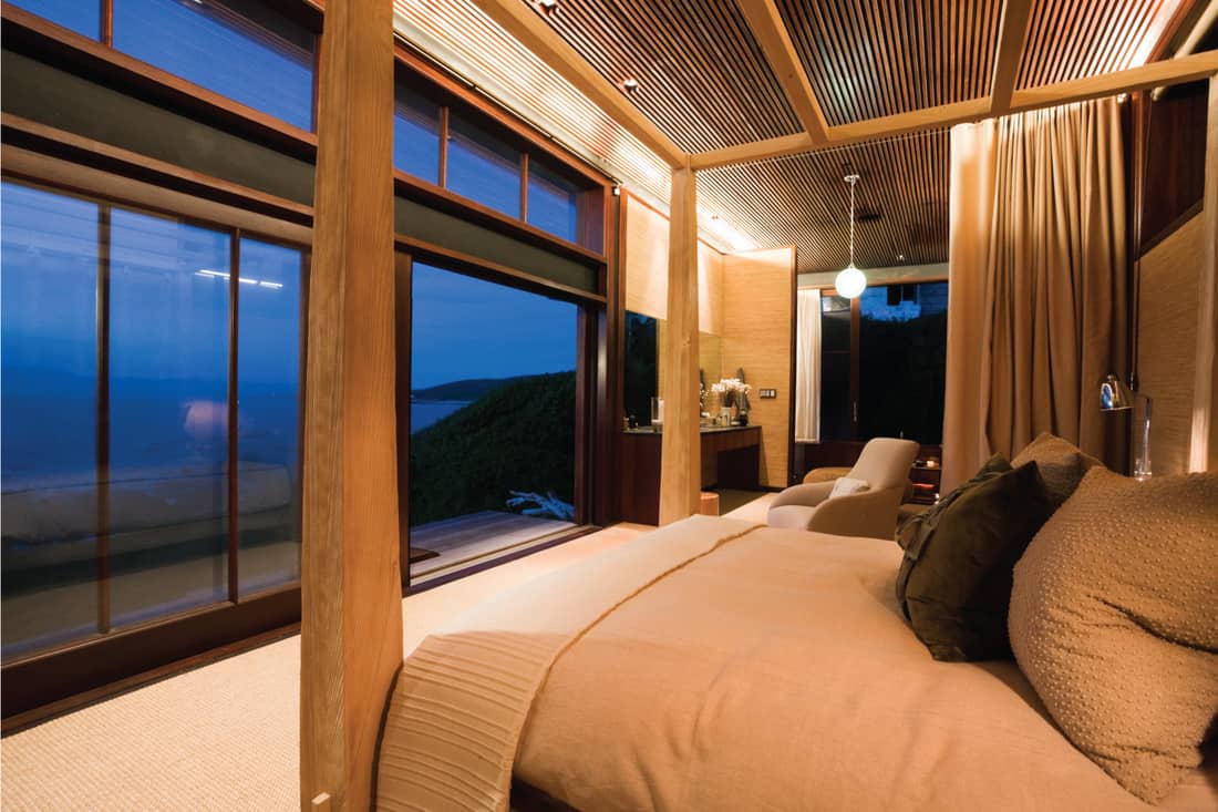 Bedroom of a luxury beach cottage with picture windows facing the sea, beige curtains around the bed. private cabana