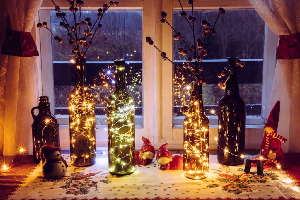 Bottles decorated with small lights and artificial red berries placed next to a window