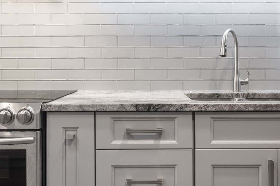 Brushed nickel faucet, stainless steel sink, granite countertop, white and grey tones. grey subway glass tile backsplash, glass top stove, stainless