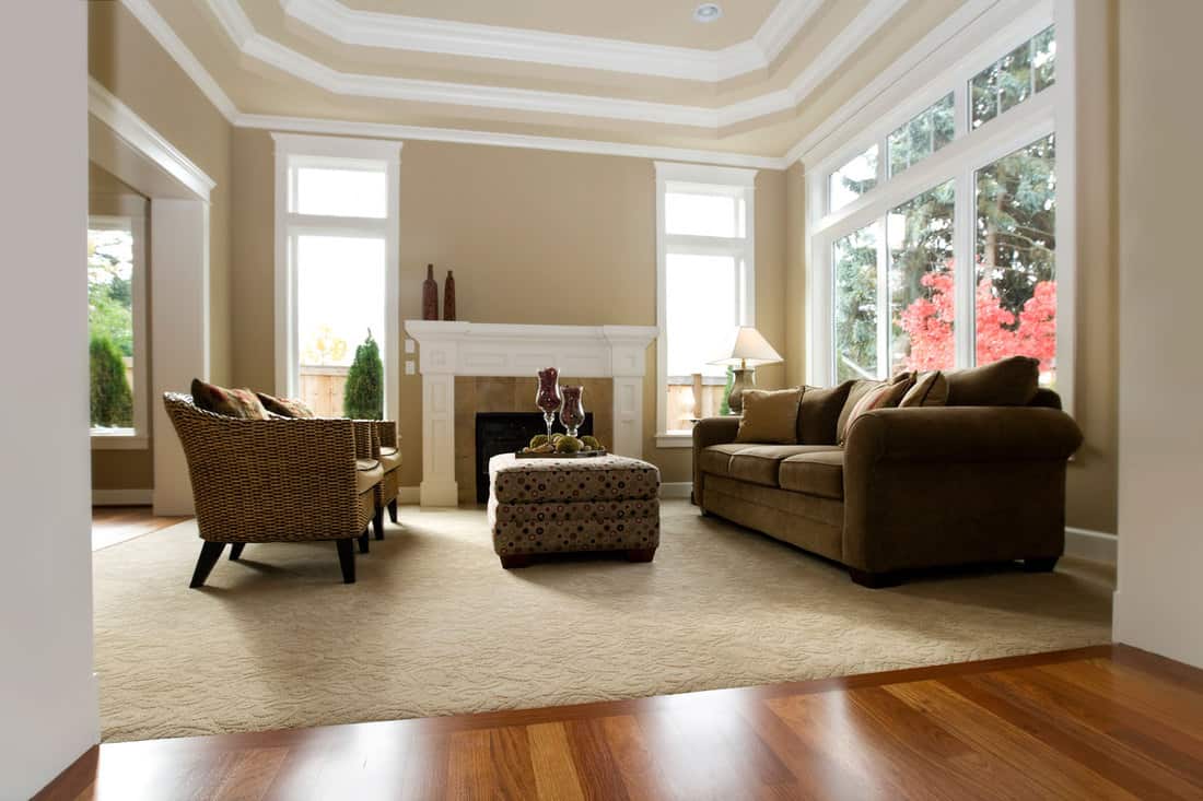 Interior of a modern living room with light brown painted walls, brown sectional sofas, brown ottoman with plants on top, and a white colored fireplace mantel, What Color Paint Goes With Beige Carpet?