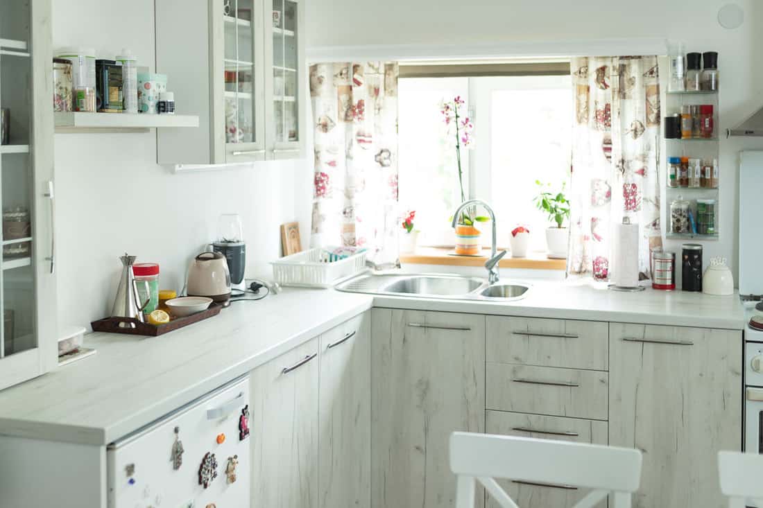 Interior of a small white themed kitchen with floral blinds on the window and indoor plants on the windowsill, What Are The Best Blinds For A Kitchen Window?