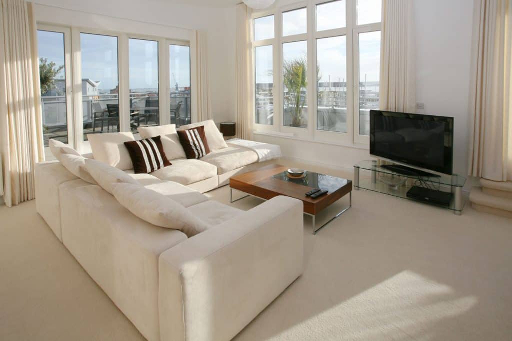 Modern interior of a gorgeous living room with white walls, beige colored curtains, white sectional sofas, and carpted flooring