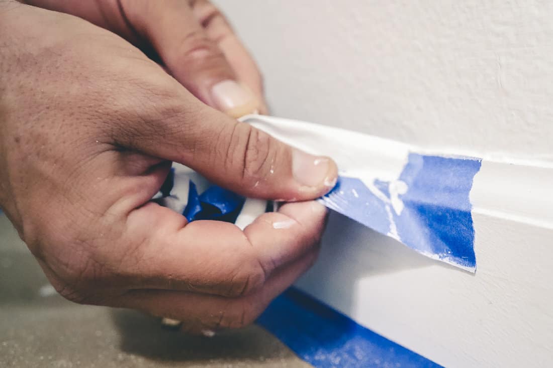 Painter pulls of blue painter's tape from the wall to reveal a clean edge baseboard