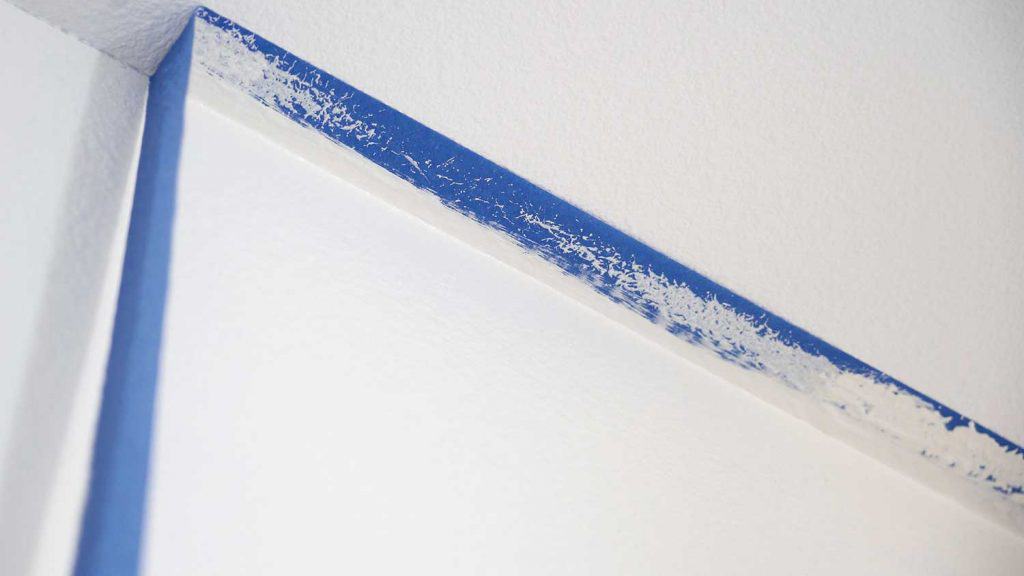 Painter pulls of blue painters tape from the wall to reveal a clean edge of the ceiling