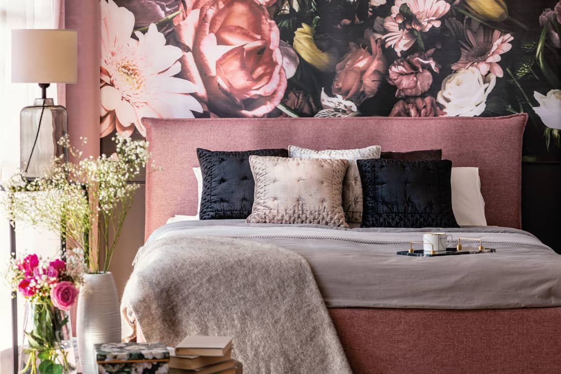 Pink and grey bed with cushions in patterned bedroom interior with flowers and lamp.