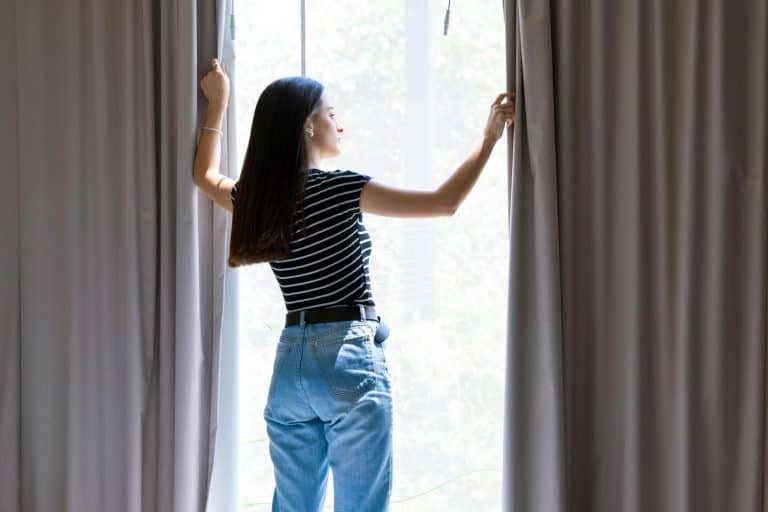 Rear view of young woman looking through window. Female is opening curtains at home, How Long Should Curtains Be For A 9 Foot Ceiling?