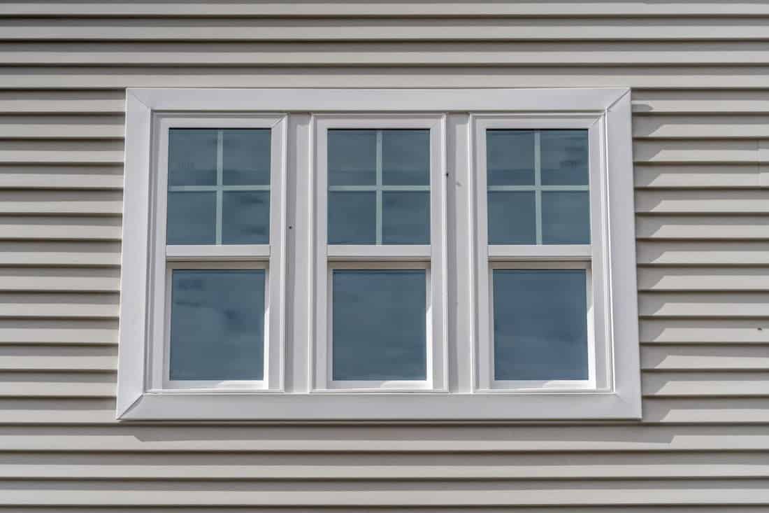Triple hung window with fixed top sash and bottom sash that slides up, sash divided by white grilles a surrounded by white elegant frame horizontal white vinyl siding on a new construction residence.