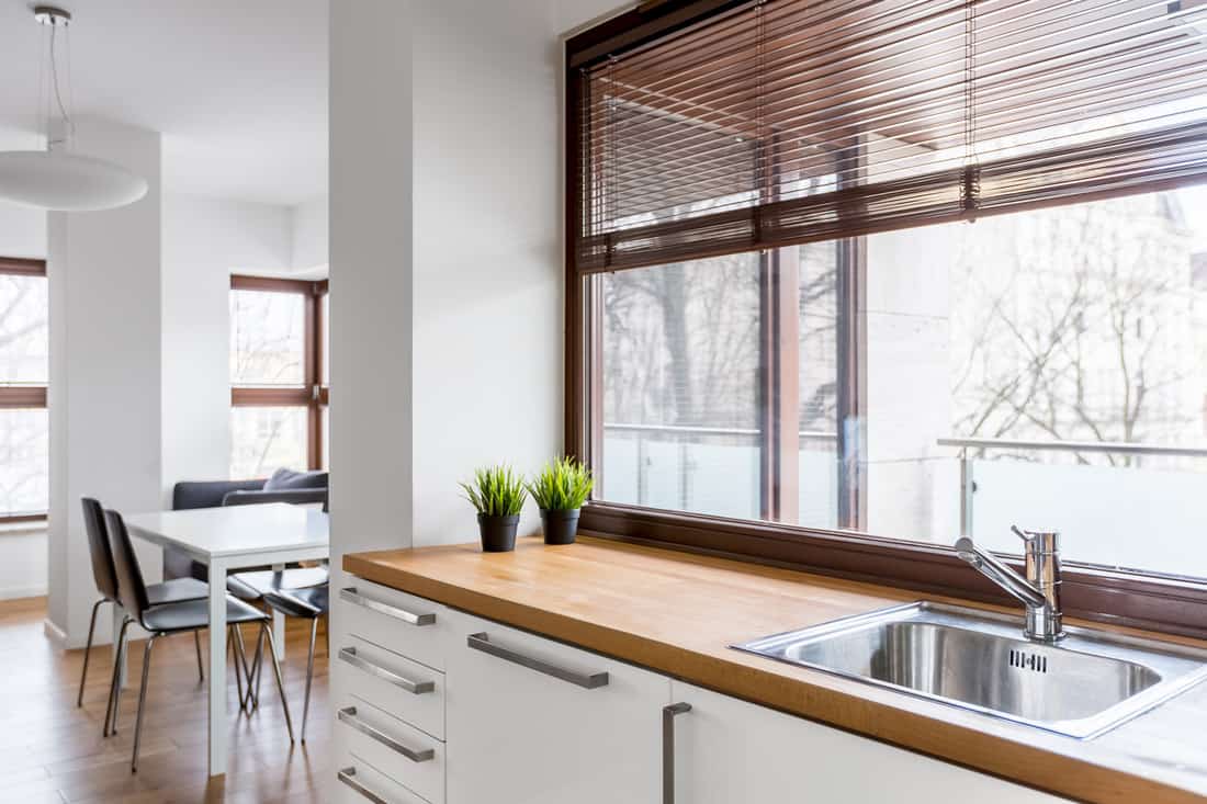 White kitchen with wooden countertop, silver sink and big window with blinds, Should Kitchen Windows Have Curtains Or Blinds?