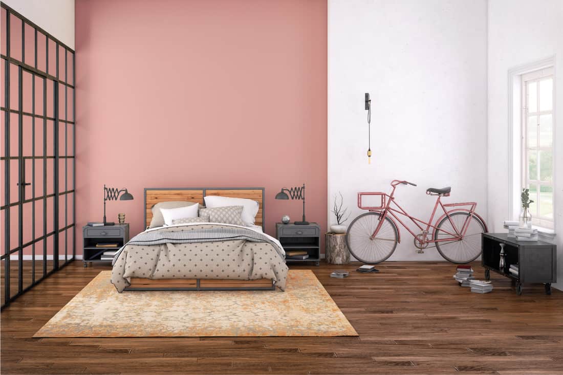 Bedroom interior, with bed, night tables, side table lamps, books on wooden floor, pink decorative bike, and white and pink wall that is rich in texture. 