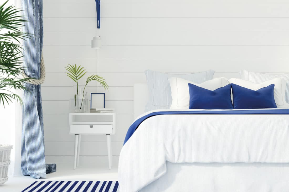Marine style bedroom with white walls, white bed frame, white nightstand, blue pillows and blanket, What Color Bedding Goes With White Furniture?