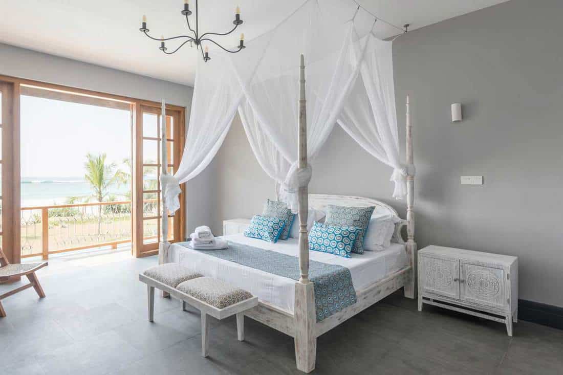 A four poster bed with mosquito net in bright hotel room with beach view on balcony, How Do You Hang Curtains Behind A Bed?
