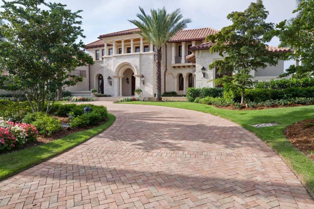 A huge luxurious mansion with a brick patterned driveway with gorgeous landscaping on the side