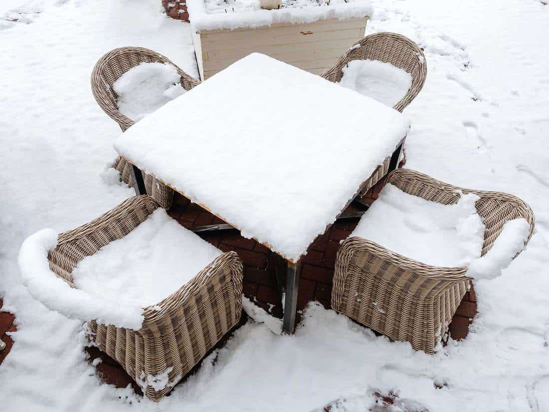 A square table with four wicker rattan chairs strewn with snow stands on a snow-covered tile