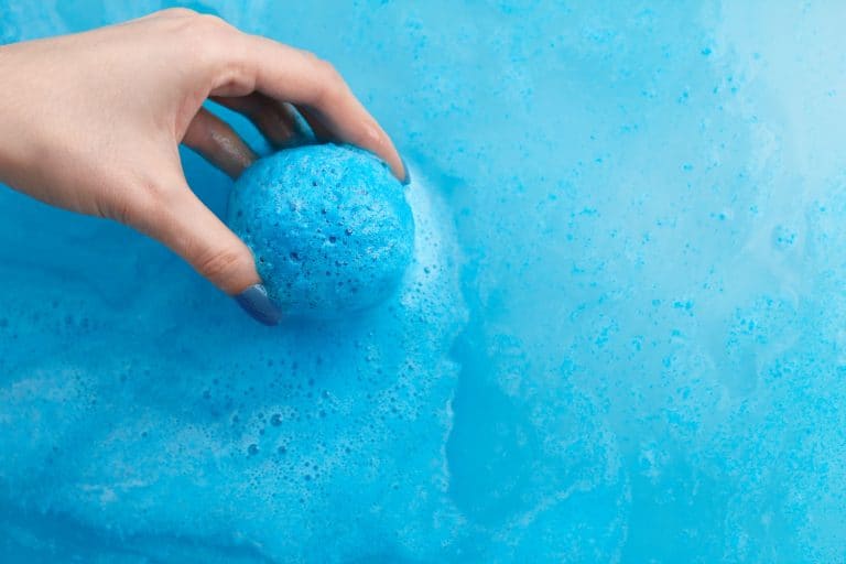 A woman putting a blue colored bath bomb on the bathtub, How To Remove Bath Bomb Stains From Bathtub [3 Excellent Options]