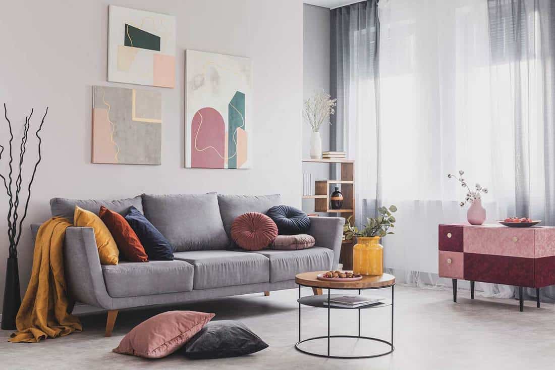 Abstract paintings hanging on white wall above a gray sofa in a living room interior with big windows