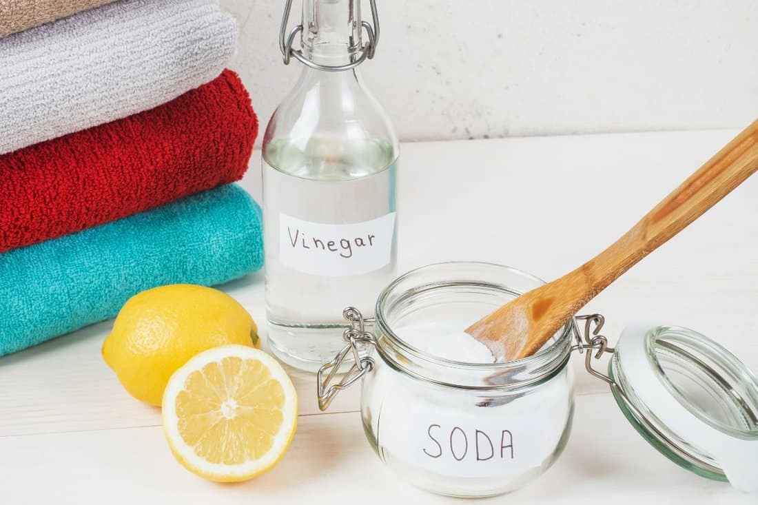 Baking soda in jar with a wooden spoon on top, vinegar, cut lemon, folded towel on a white background. The concept of organic removing stains on clothes