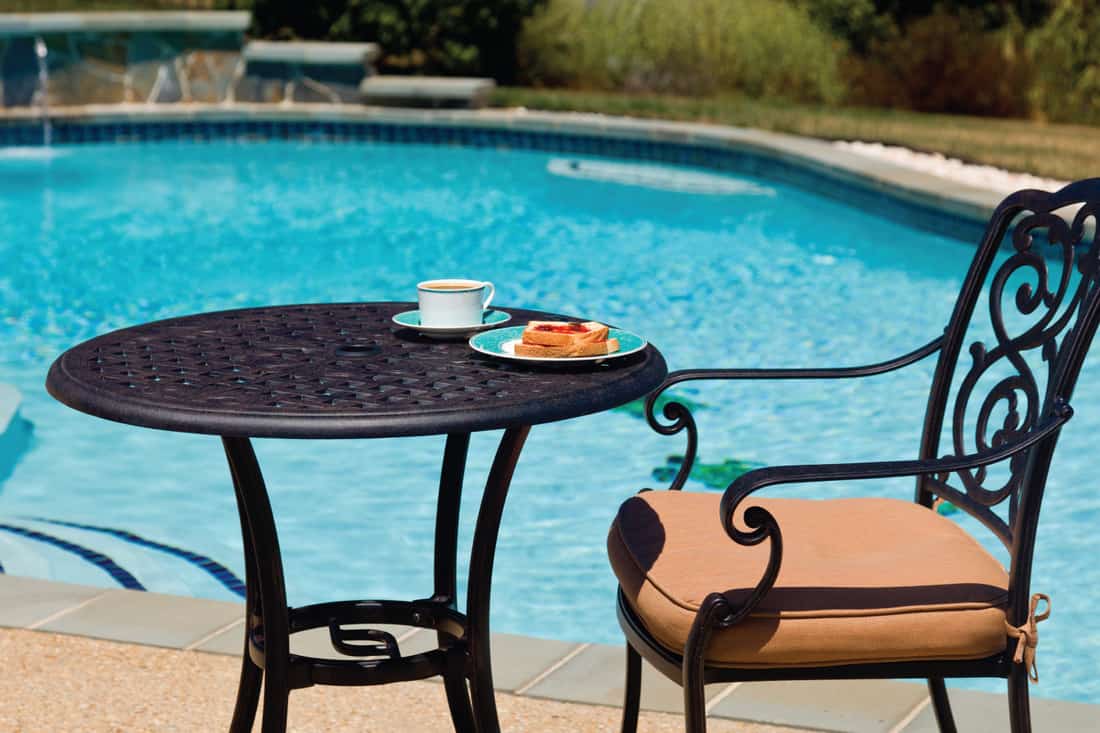 Coffee and plate on cast aluminum table and single chair by the side of swimming pool in back yard