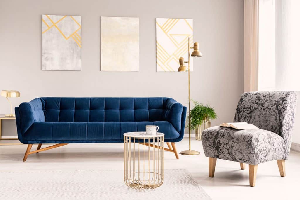 Interior of a boho themed living room with a long blue colored sofa, gold framed floor lamp, light gray colored wall, and patterned black and white accent chair