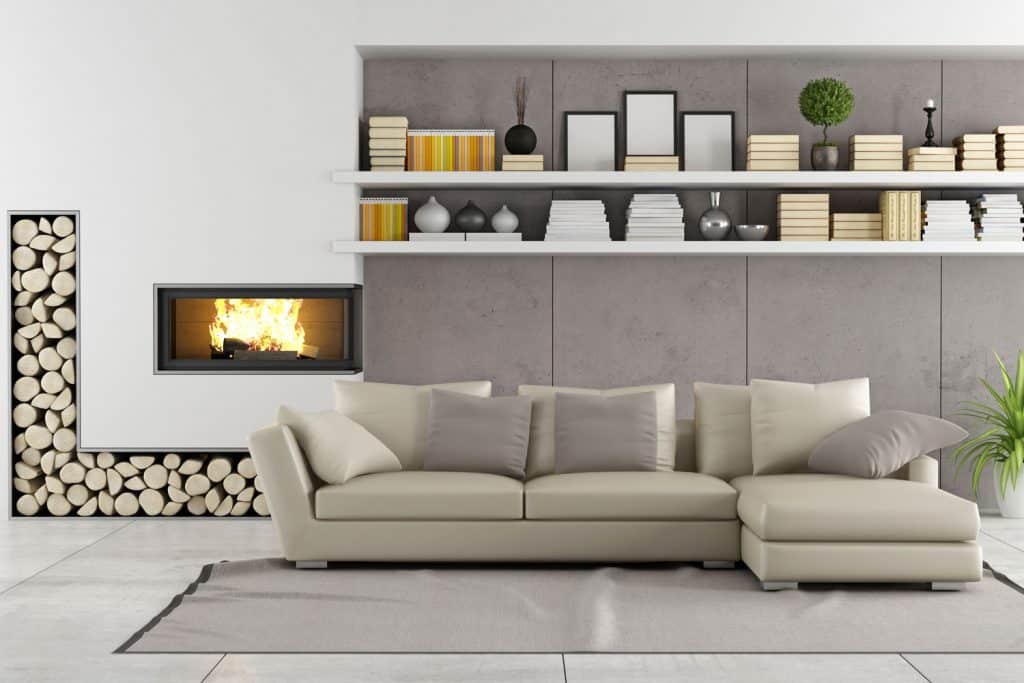 Interior of a modern contemporary living room with a light dark-white colored sectional sofa, office supplies on the bookshelves, and a fireplace with a firewood design