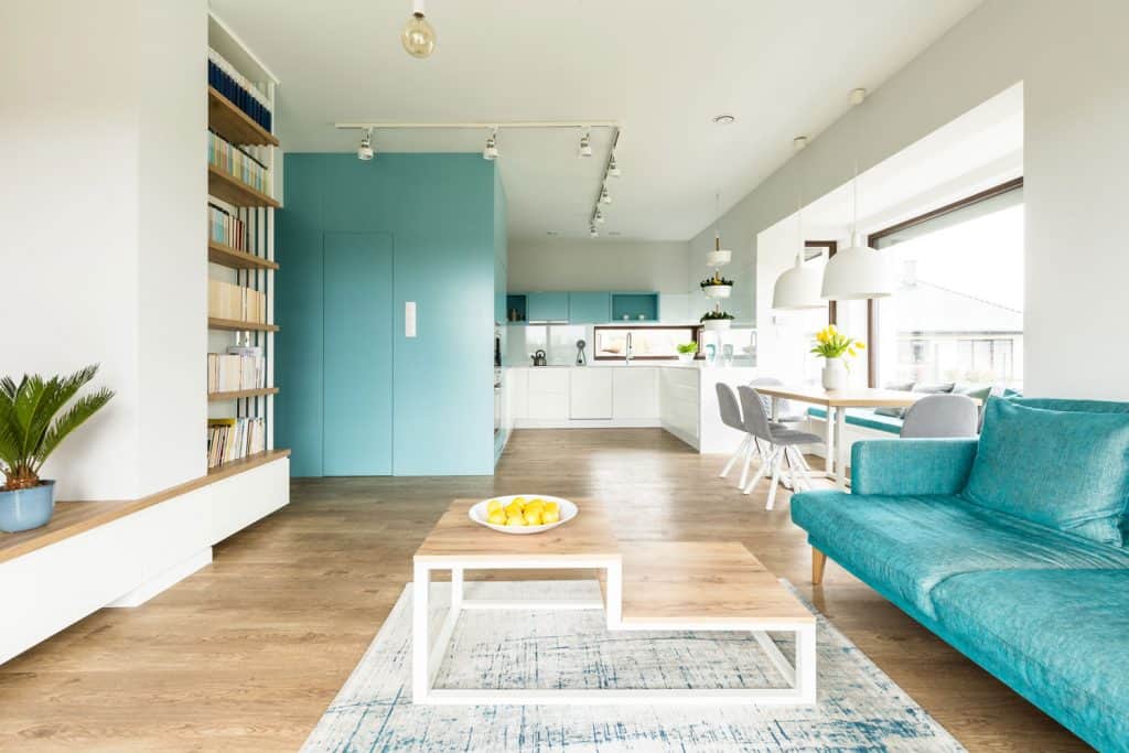 Interior of an ultra modern living room with wooden laminated flooring, a long sky blue sofa, and kitchen cabinets painted in sky blue