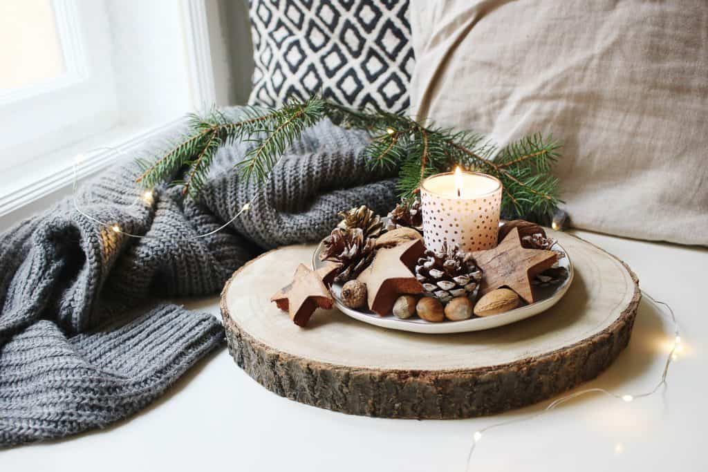 Woody scented candle, Burning candle decorated by wooden stars, hazelnuts and pine cones standing near window on wooden cut board.