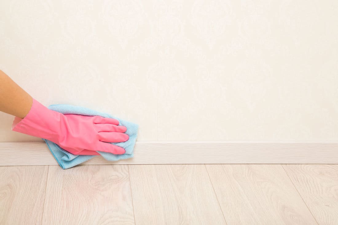 Young adult woman hand in pink rubber protective glove using blue dry rag and wiping light wooden baseboard surface in room at home. Closeup. Empty place for text on wall background. 