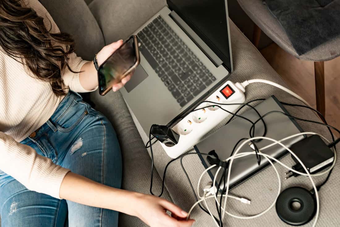 Jeune femme sur un canapé chargeant plusieurs appareils technologiques avec prise électrique, Comment cacher les cordons électriques dans le salon ?