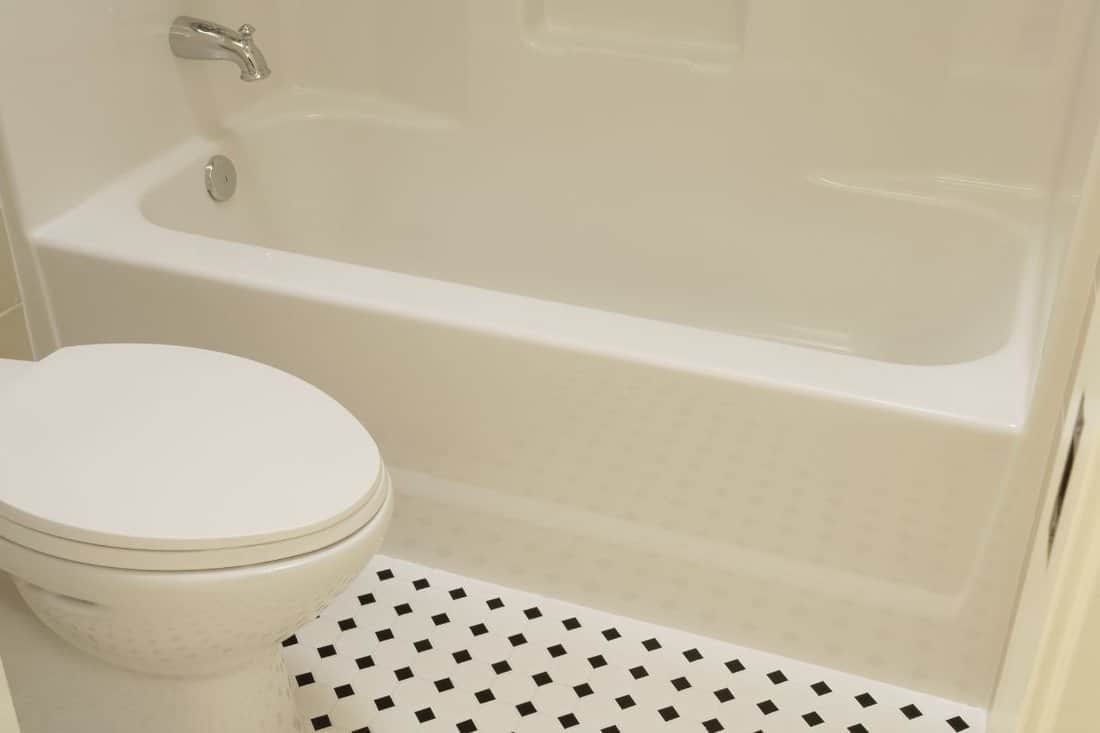A white acrylic bathtub and porcelain toilet of a bathroom are sitting on a diamond patterned vinyl flooring in a bathroom. Horizontal shot.