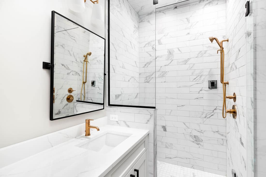 An all white master bathroom with gold hardware and faucets showing the vanity and shower with the door open. The shower has glass walls/door and beautiful tiles.