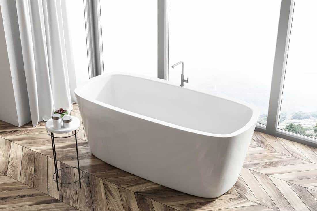 Bathroom interior with a wooden floor, a panoramic window with a mountain view and a white angular bathtub