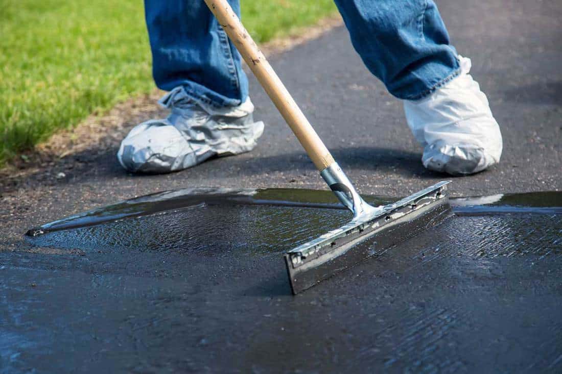 Close-up photo of a home owner seal coating their driveway, Does Driveway Sealer Fill Cracks? [And Which One To Use!]