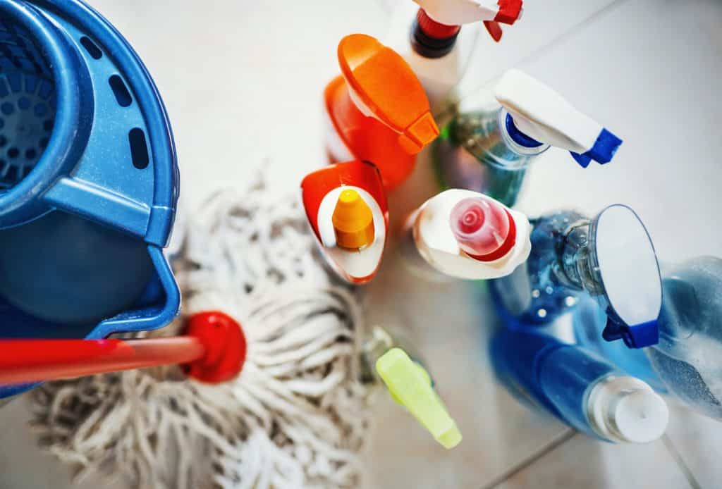 Vue de dessus en gros plan de produits de nettoyage domestiques méconnaissables avec un seau bleu et une vadrouille sur le côté.  Tous les produits placés sur le carrelage blanc de la salle de bain.