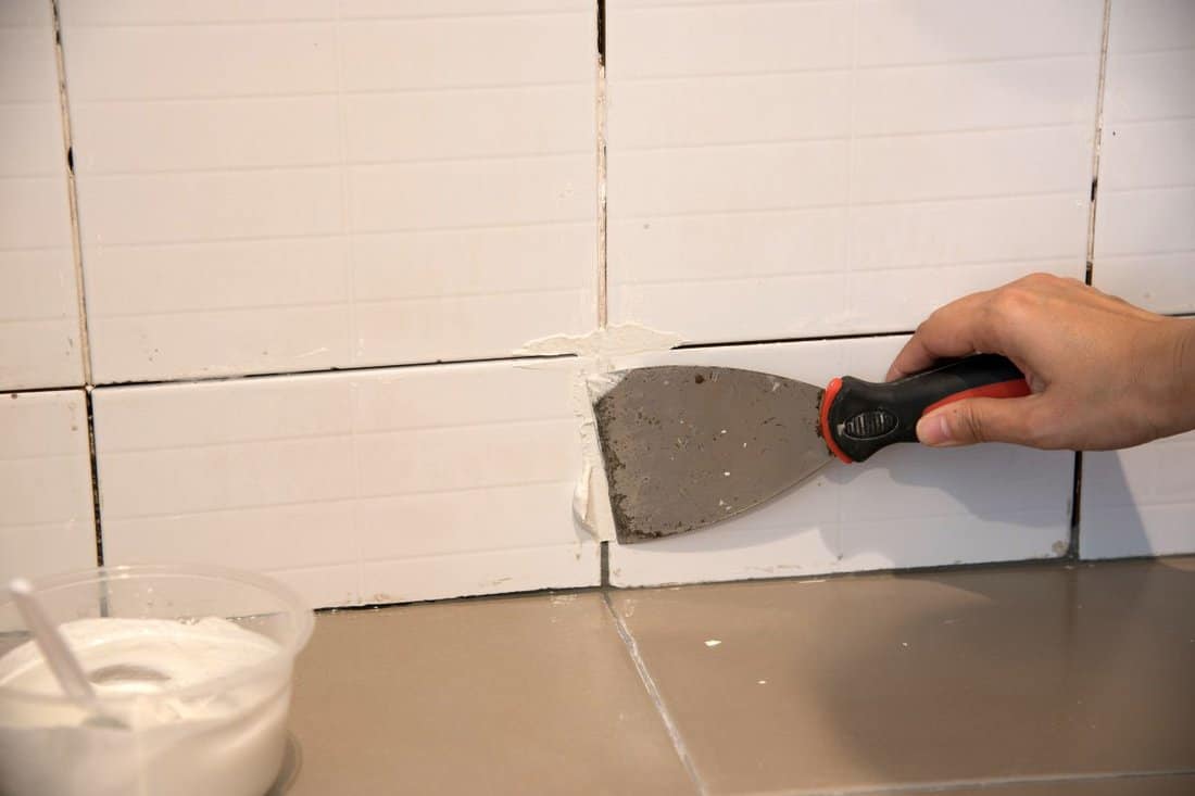 DIY.The worker repairing tiler on the wall with trowel, removes the glue residues from the interstice seam and install new glue or white silicone, the technology of laying tiles and finishing.