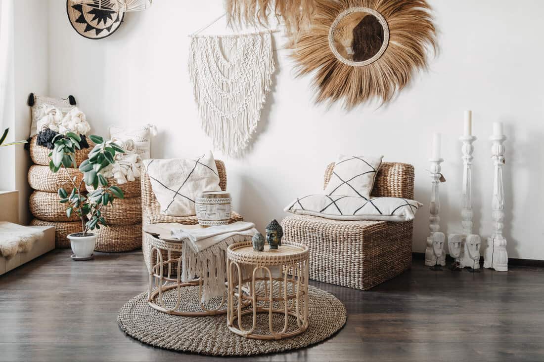 Home decor concept. Comfortable wicker furniture, rattan armchair with cushions, bamboo coffee table and macrame on white wall in cozy living room with ethnic interior design 