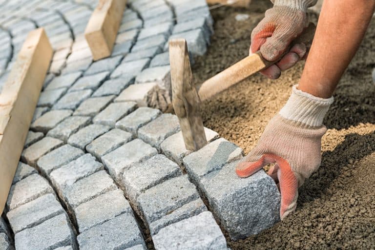 Laying paving stone, just hands in gloves and a hammer in action, hammering on the paving stones, How To Seal Pavers With Sand [6 Easy Steps To Follow!]
