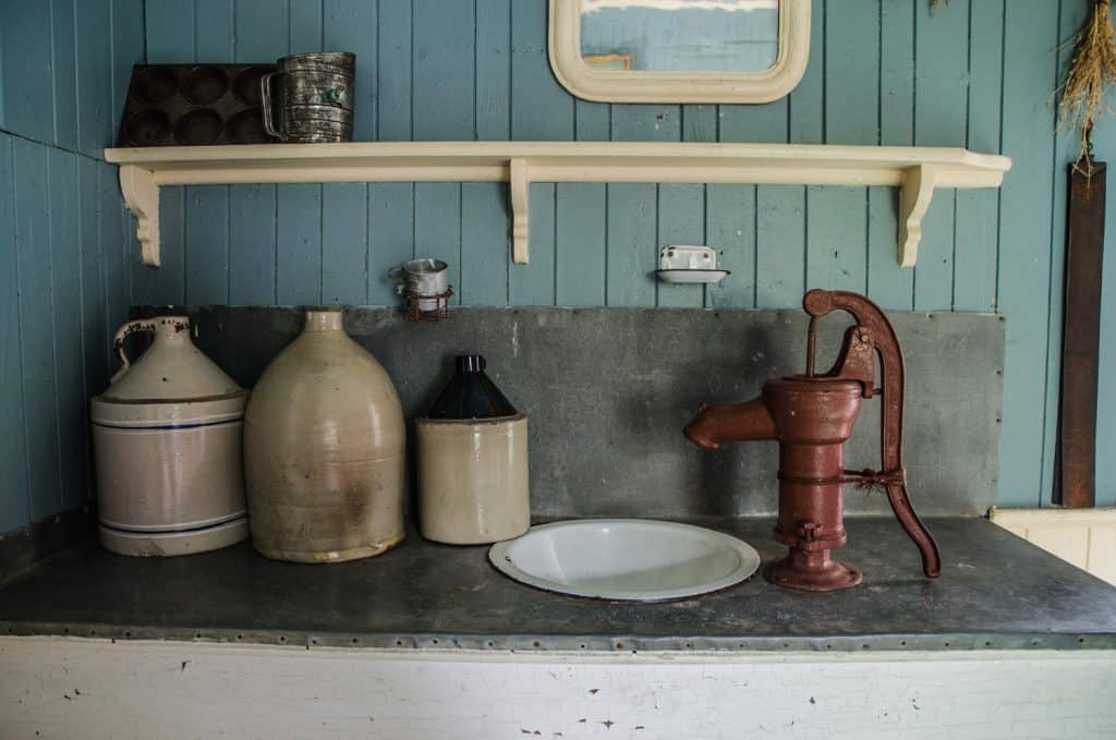 Old and antique kitchen counter with old jugs, water pump, sink, mirror and shelf above the counter