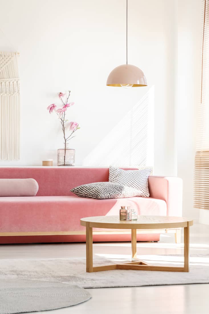 Retro style, millennial pink pendant lamp above a simple, wooden coffee table in a sunny, white living room interior with patterned pillows on a velvet sofa