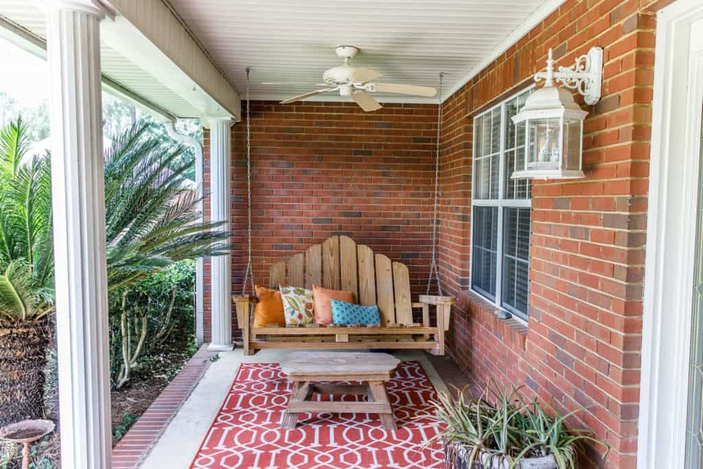 Rustic inspired front porch of a house with brick walls, wooden two seater swing, and a white beadboard ceiling