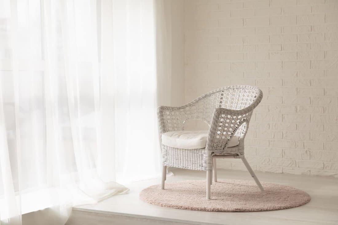 White wicker armchair standing in empty modern room with big windows, curtains and soft carpet rug, elegant and quiet cozy interior in daylight, decorative