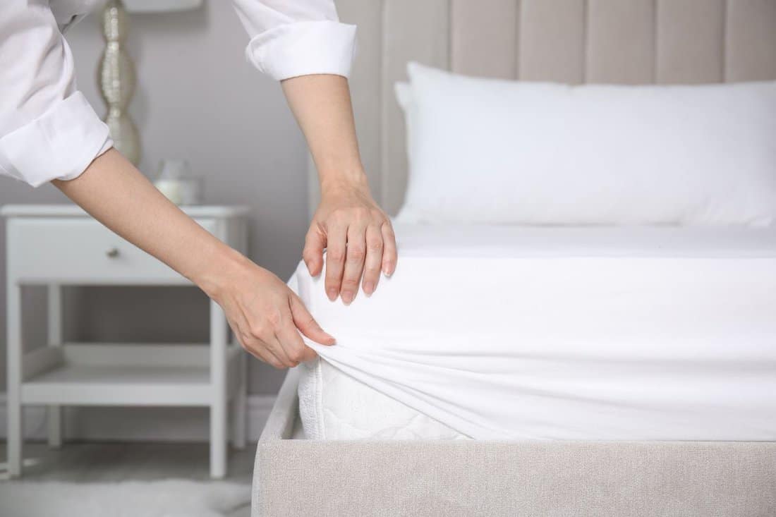 Woman putting white fitted sheet over mattress on bed indoors,