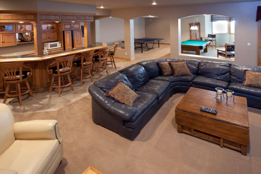 A large and spacious rustic inspired basement flooring with a large black leather sectional sofa, wooden themed bar, and carpeted flooring