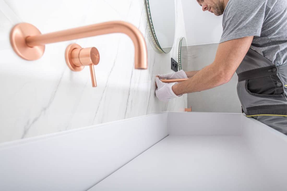 Caucasian Handyman Worker Fastening By Hand Copper Color Faucet To Marble Tile Wall In Bathroom.