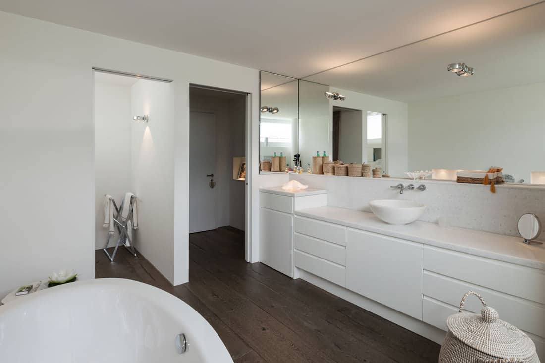 Elegant and white inspired bathroom with white painted cabinets, long span mirror, and a huge white bathtub, How Tall Are Bathroom Cabinets? [Standard Dimensions Explored]