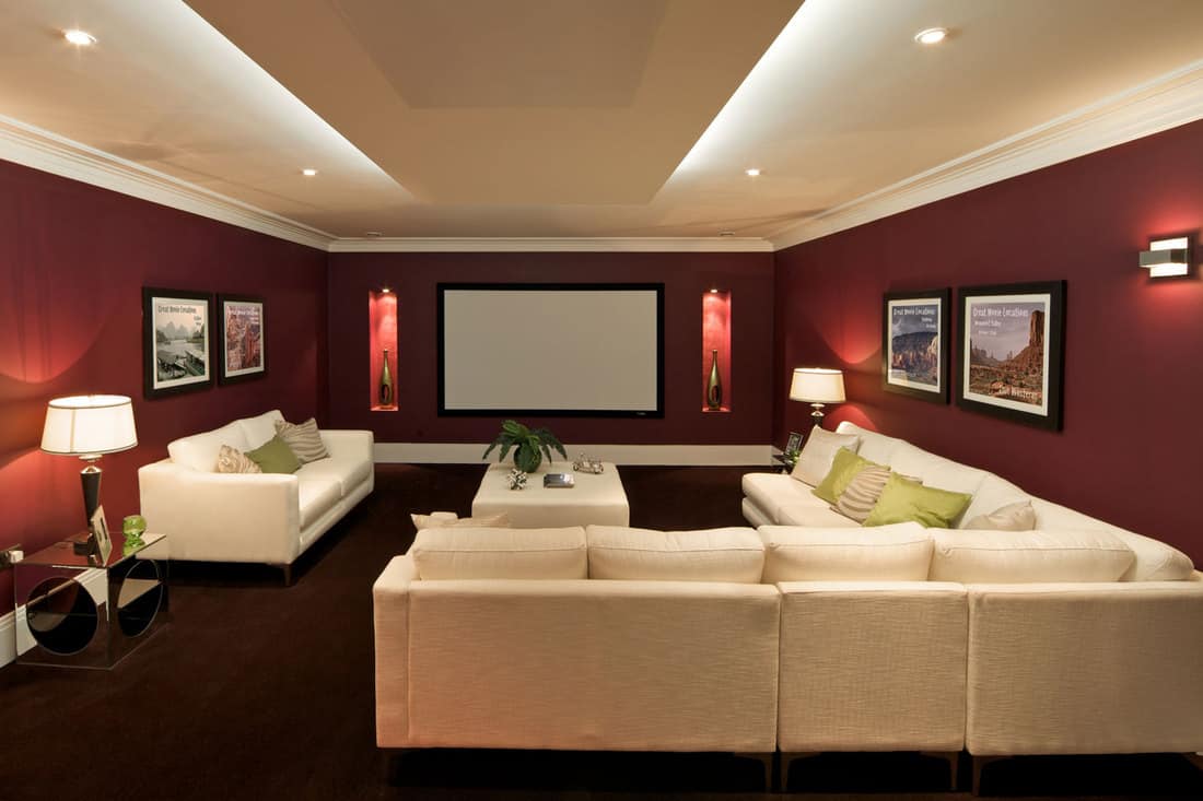 Interior of a contemporary basement with a white ceiling, red walls with huge landscape picture frames, and a huge beige colored sectional sofa, What Is The Best Color For A Dark Basement?