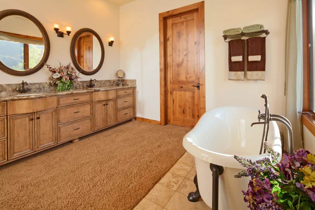 Interior of an elegant and rustic bathroom with wooden door cabinetry, carpeted flooring, and two round mirrors