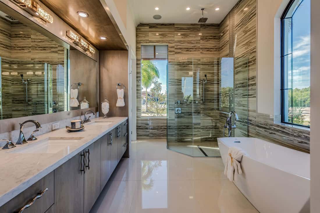 Interior of an ultra modern bathroom with wooden cladding on the walls, huge mirror on the vanity, glass shower wall, and a huge window near the bathtub, What Wall Color Goes With Gray Bathroom Cabinets?