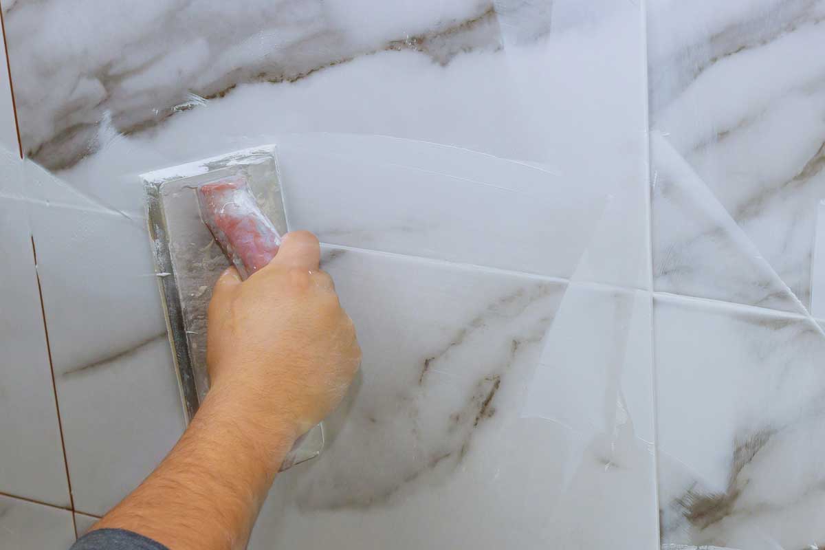 Male hand with the rubber applies grout on a seam between tiles, How Long Does Bathroom Grout Last?