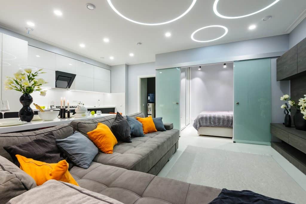 An ultra modern and luxurious apartment with a huge gray sectional sofa, bright colored throw pillows, and recessed lighting, Can Vs Canless Recessed Lighting - What Are The Differences?