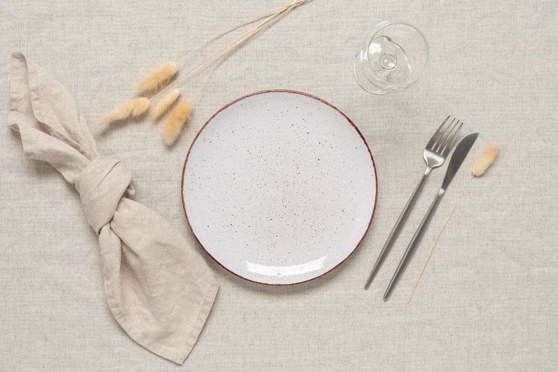 Autumn table setting. A white plate, cutlery and dry grass lagurus on a beige linen textile background. Top view, flat lay. Selective focus.